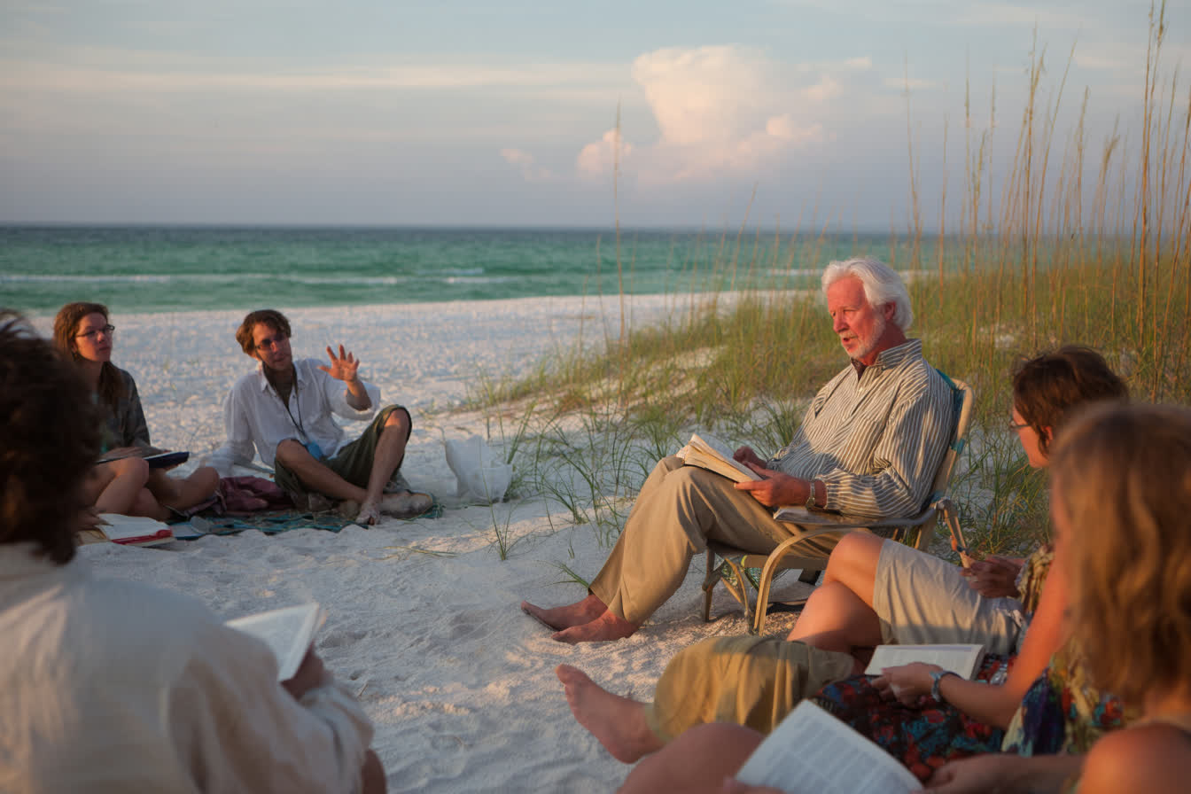 A recitation on the beach: a professor in a folding chair reads to students seated on the sand, sea oats and the Gulf behind them